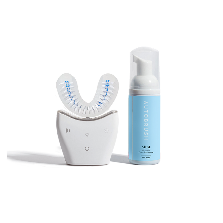 White U-shaped Autobrush device with built-in bristles, shown beside a blue bottle of mint fluoride foaming toothpaste.
